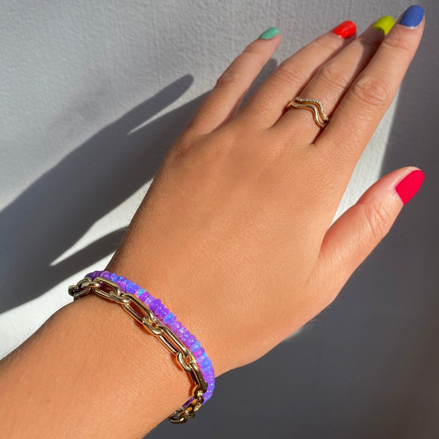 Shimmering beaded bracelet made of smooth opals in shades of bright purple on a gold linking lobster clasp. Styled on a wrist with a diamond cut link chain bracelet.