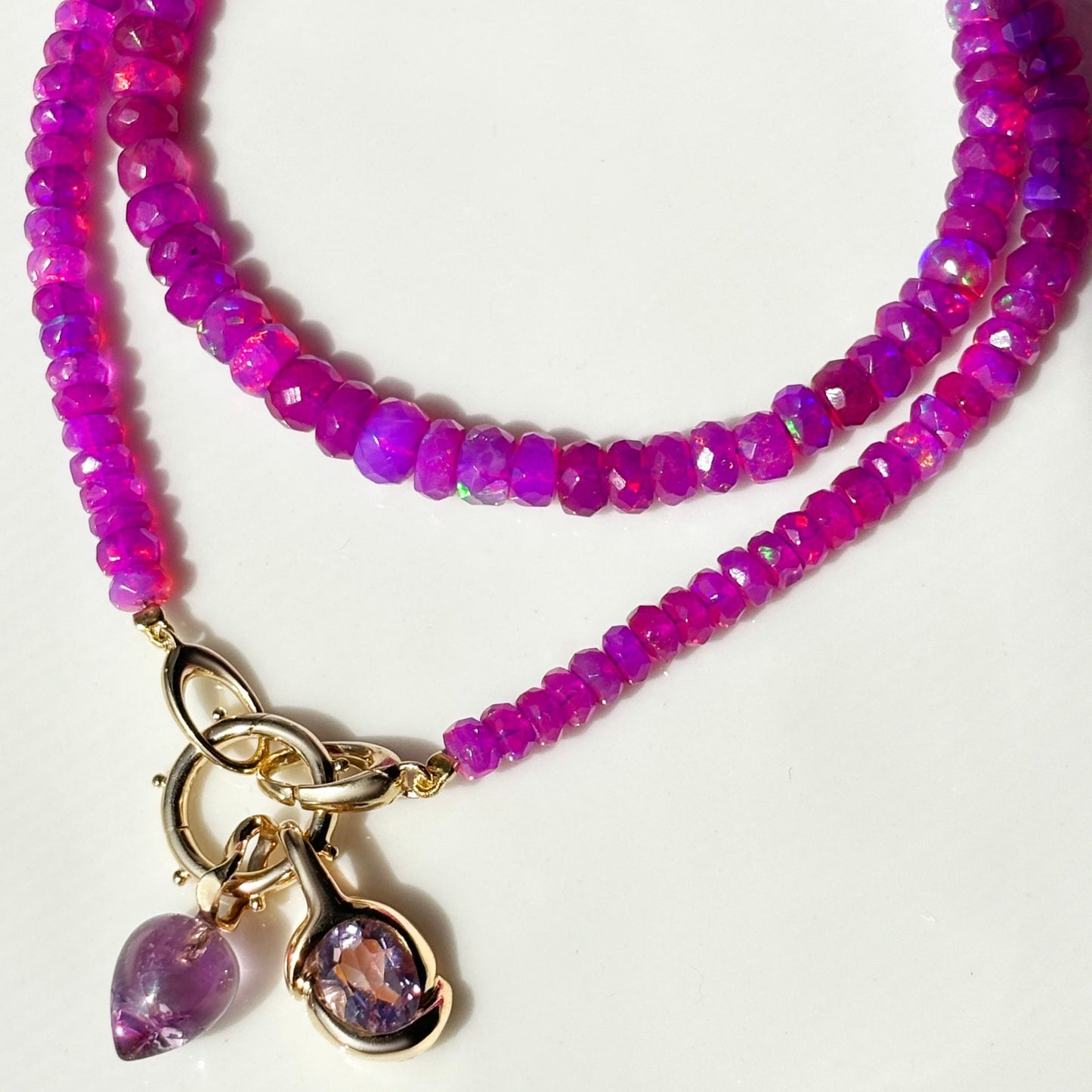 Shimmering beaded necklace made of faceted opals in shades of fuchsia pink on a gold linking ovals clasp. Styled on a neck layered with the beaded round charm lock, amethysts molten knot charm and acorn charm.