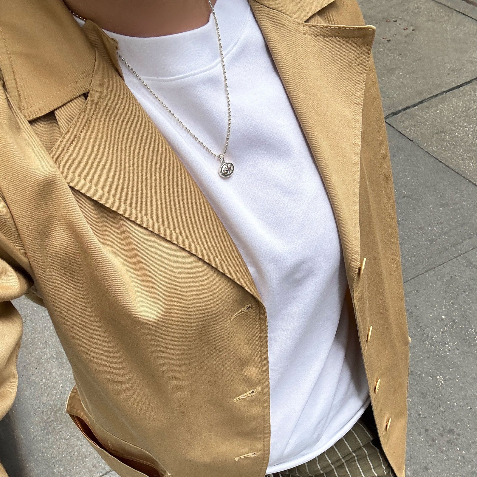 White gold bead chain with molten solitaire charm with oval diamond over a white tee, striped pants, and a camel jacket.