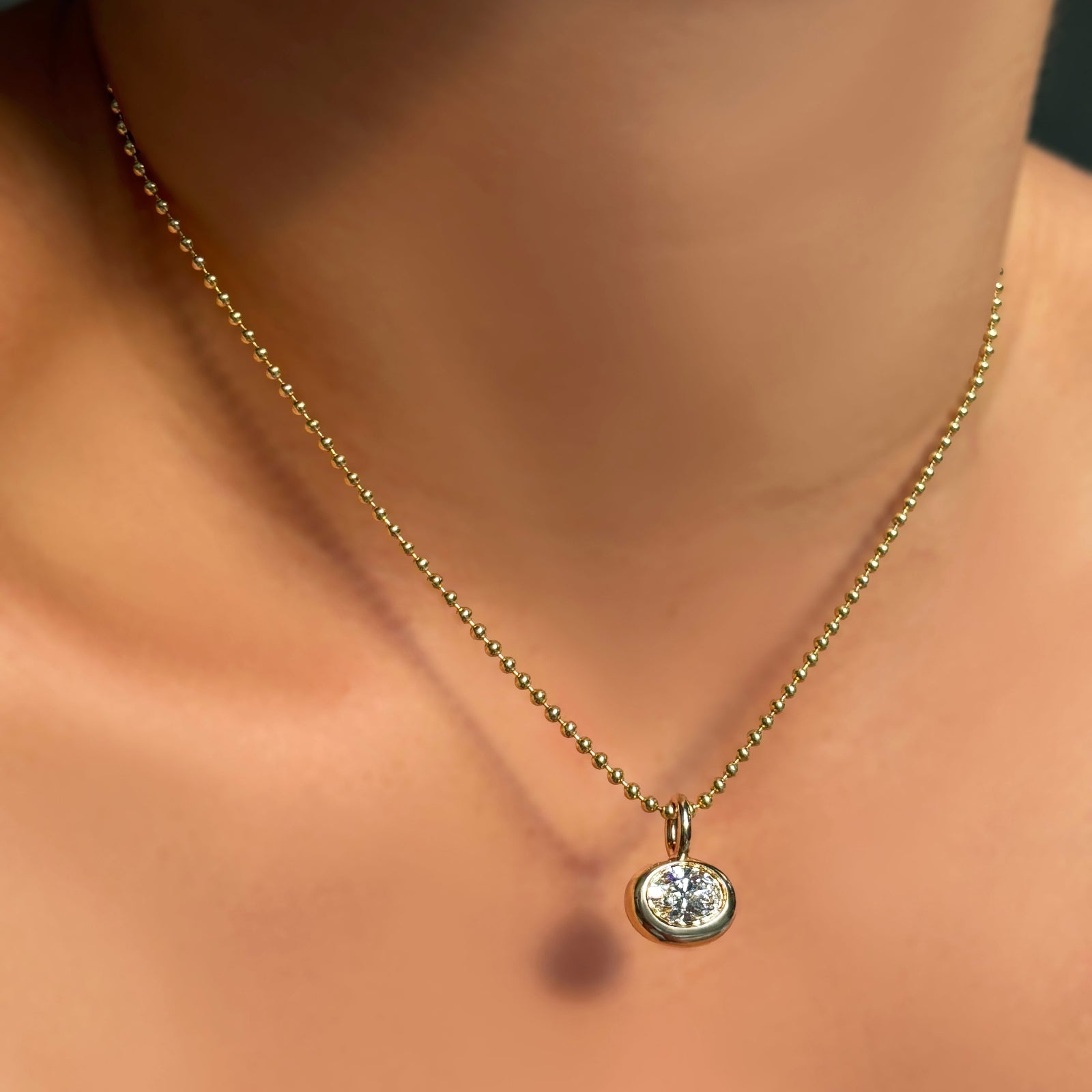 2mm bead chain necklace and small oval molten solitaire charm in 14k yellow gold on a model's neck.