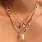 A model wears a bead chain from which hangs a quill bezel charm with a diamond, a 6mm irregular cable chain necklace and a mother of pearl drop charm, all of these, in yellow gold.