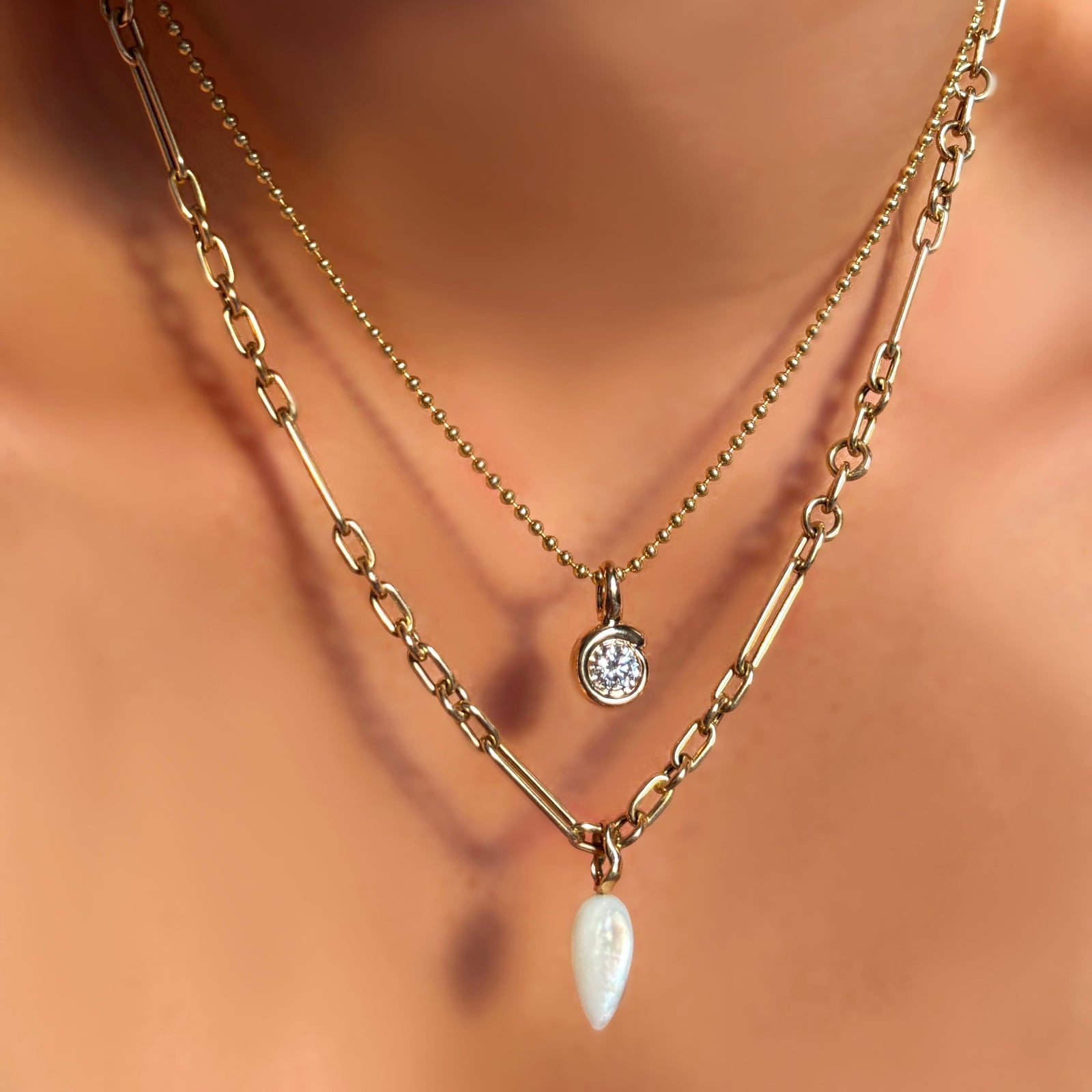 A model wears a bead chain from which hangs a quill bezel charm with a diamond, a 6mm irregular cable chain necklace and a mother of pearl drop charm, all of these, in yellow gold.