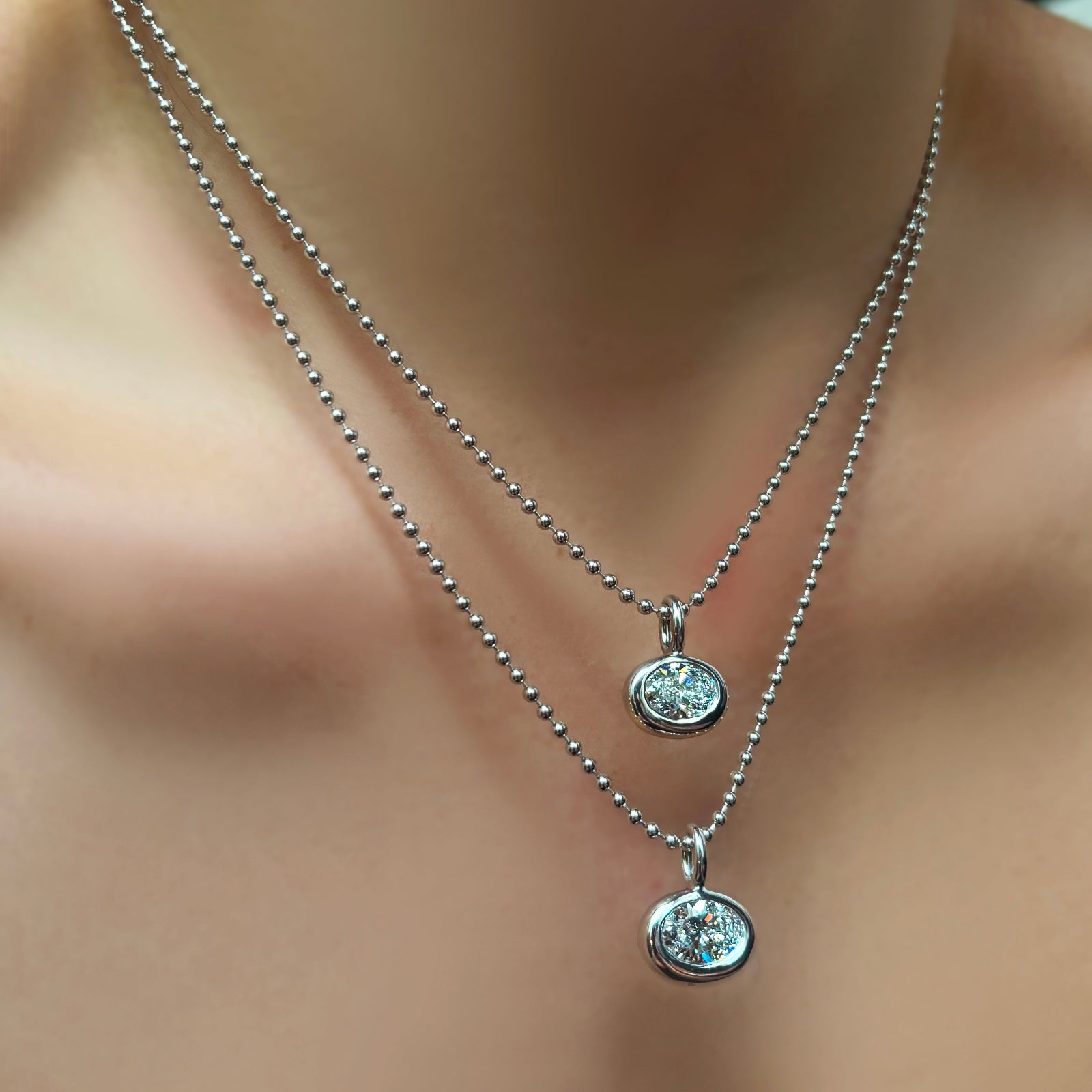 Small and regular molten solitaire charms with oval diamonds in white gold on 2mm bead chains on a model's neck.