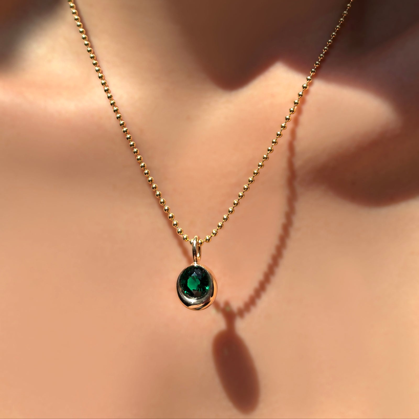 14k gold 2mm bead chain with a molten solitaire charm with round emerald on a model's neck.