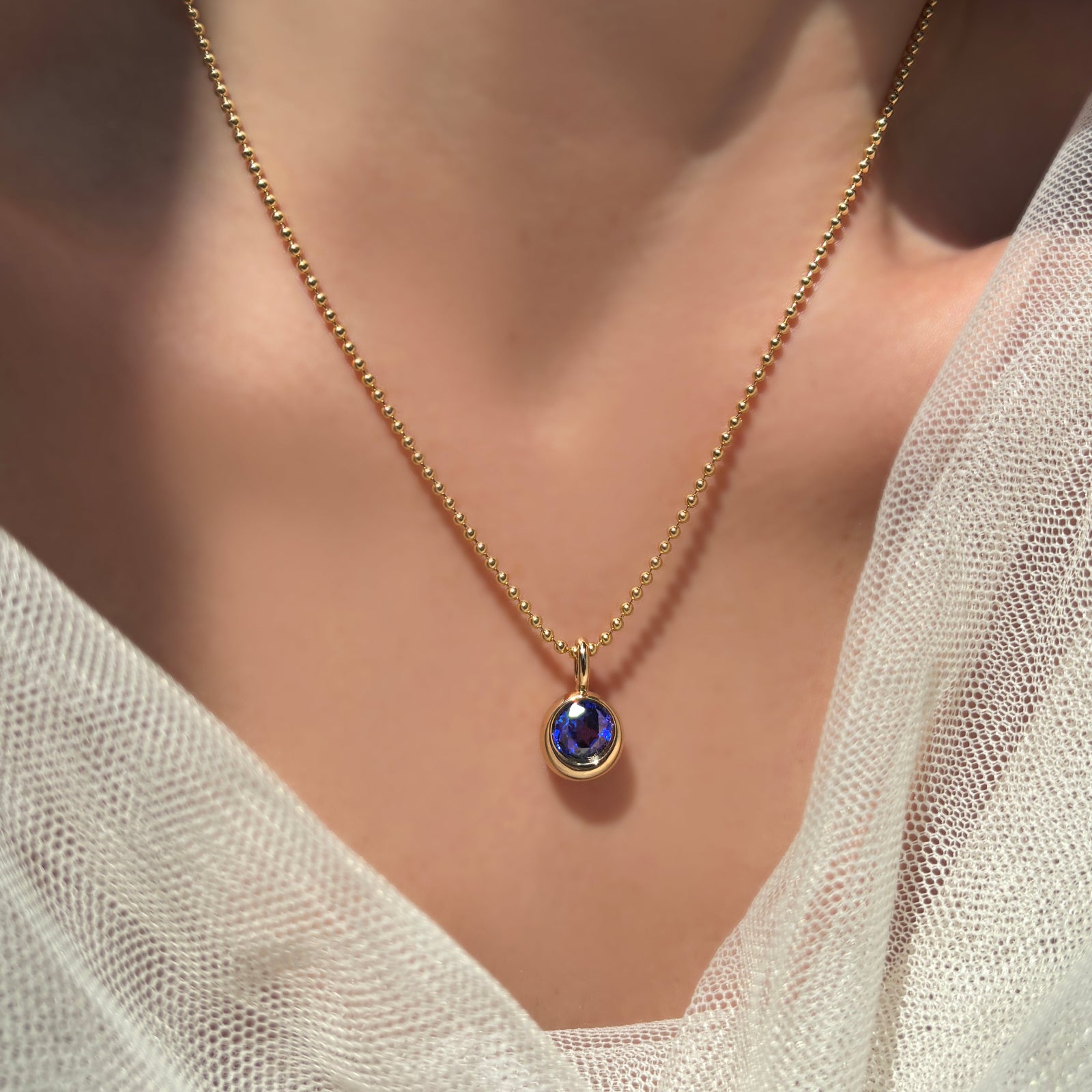 2mm bead chain in 14k yellow gold with a molten solitaire charm with round sapphire on a model's neck with some bridal tulle surrounding it.