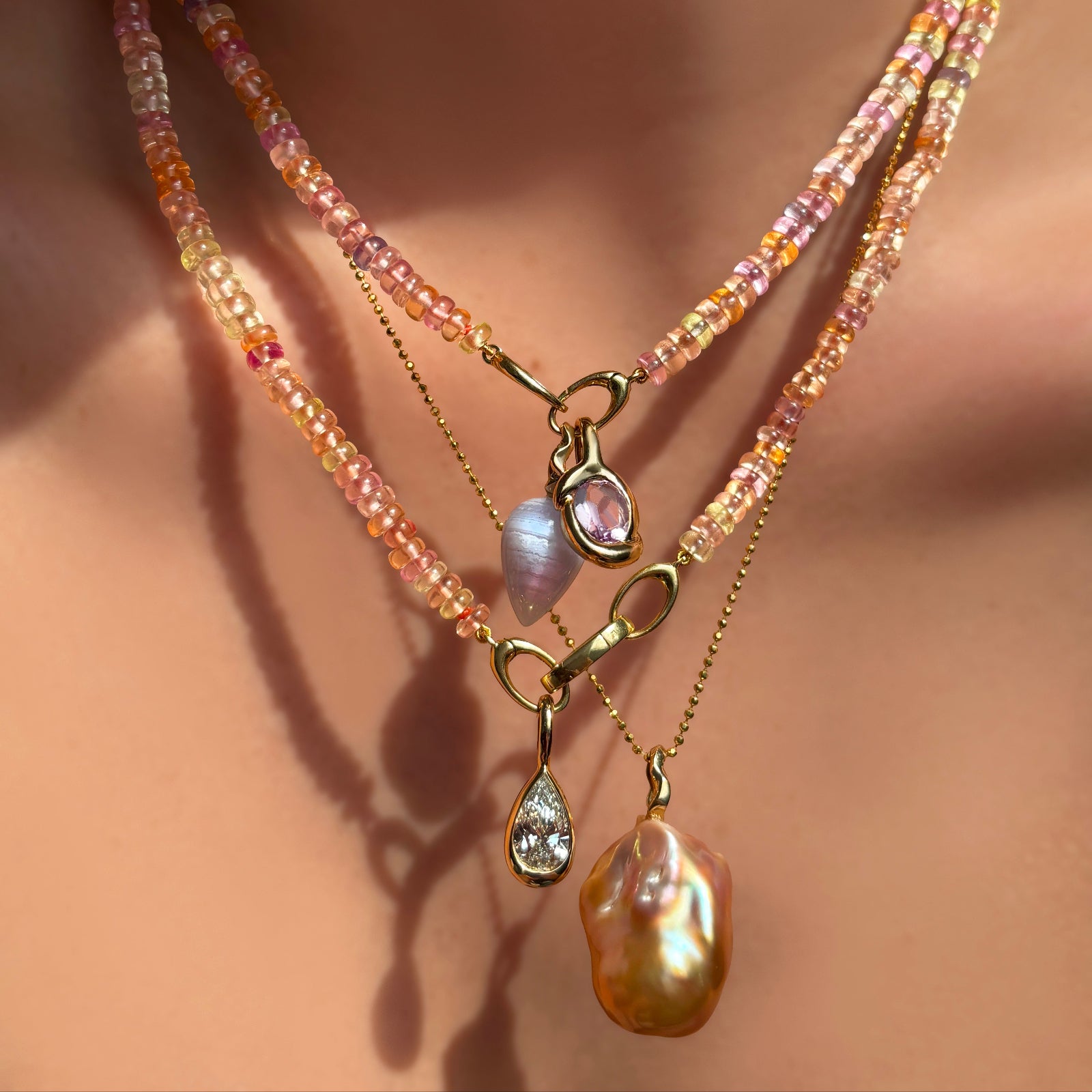 Blush sapphire necklaces layered with matching gemstone, diamond, and baroque pearl charms on a model's neck.