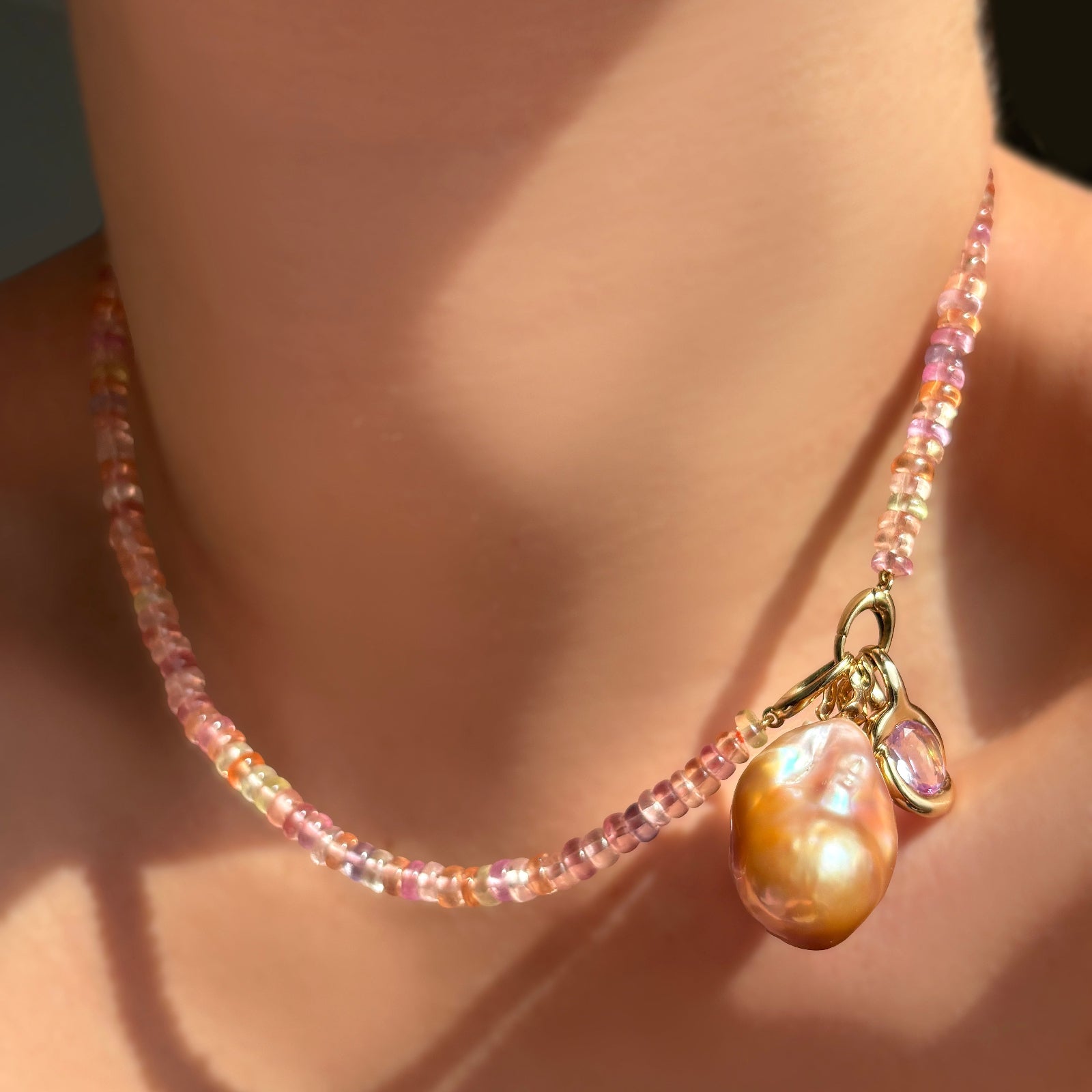 Blush sapphire necklace with its linking ovals clasp turned to the front, holding a blush baroque pearl and molten knot charm with amethyst on a model's neck.