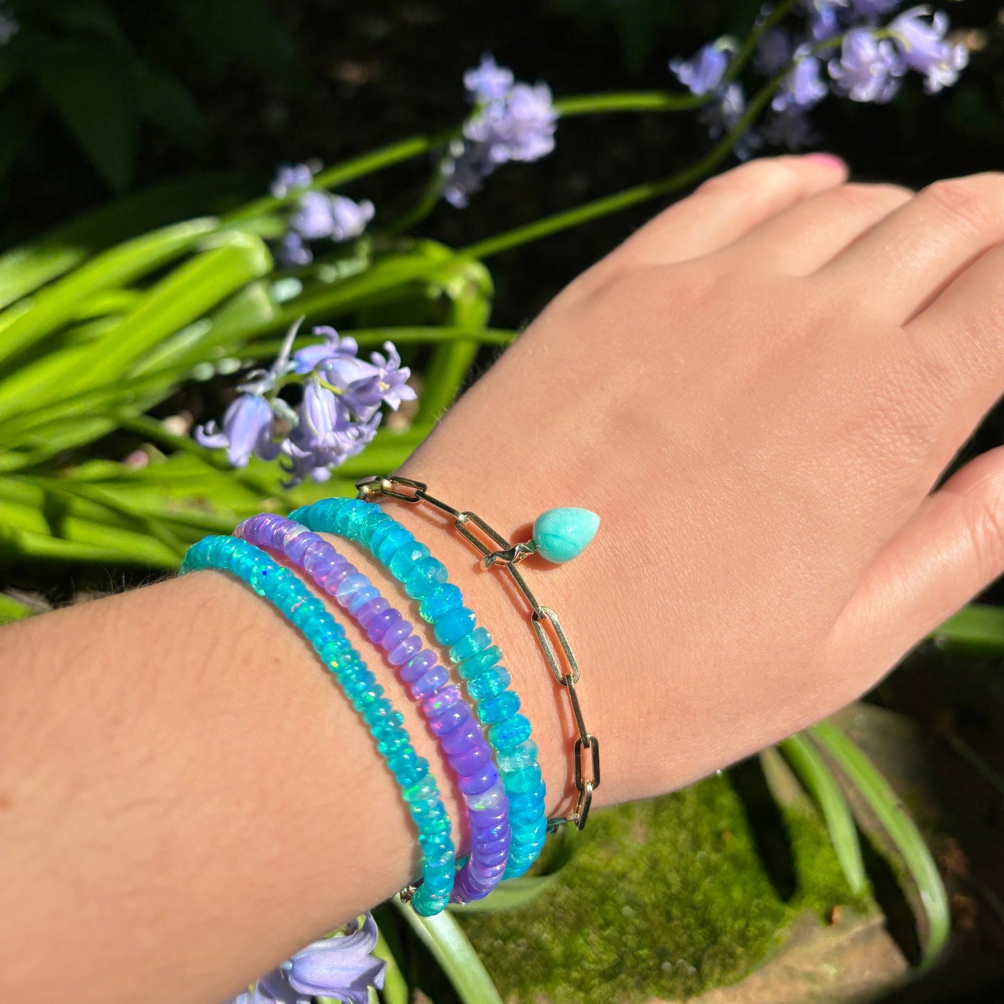 Shimmering beaded bracelet made of faceted opals in shades of bright blue on a gold linking lobster clasp. Styled on a wrist with a chain and beaded bracelets 