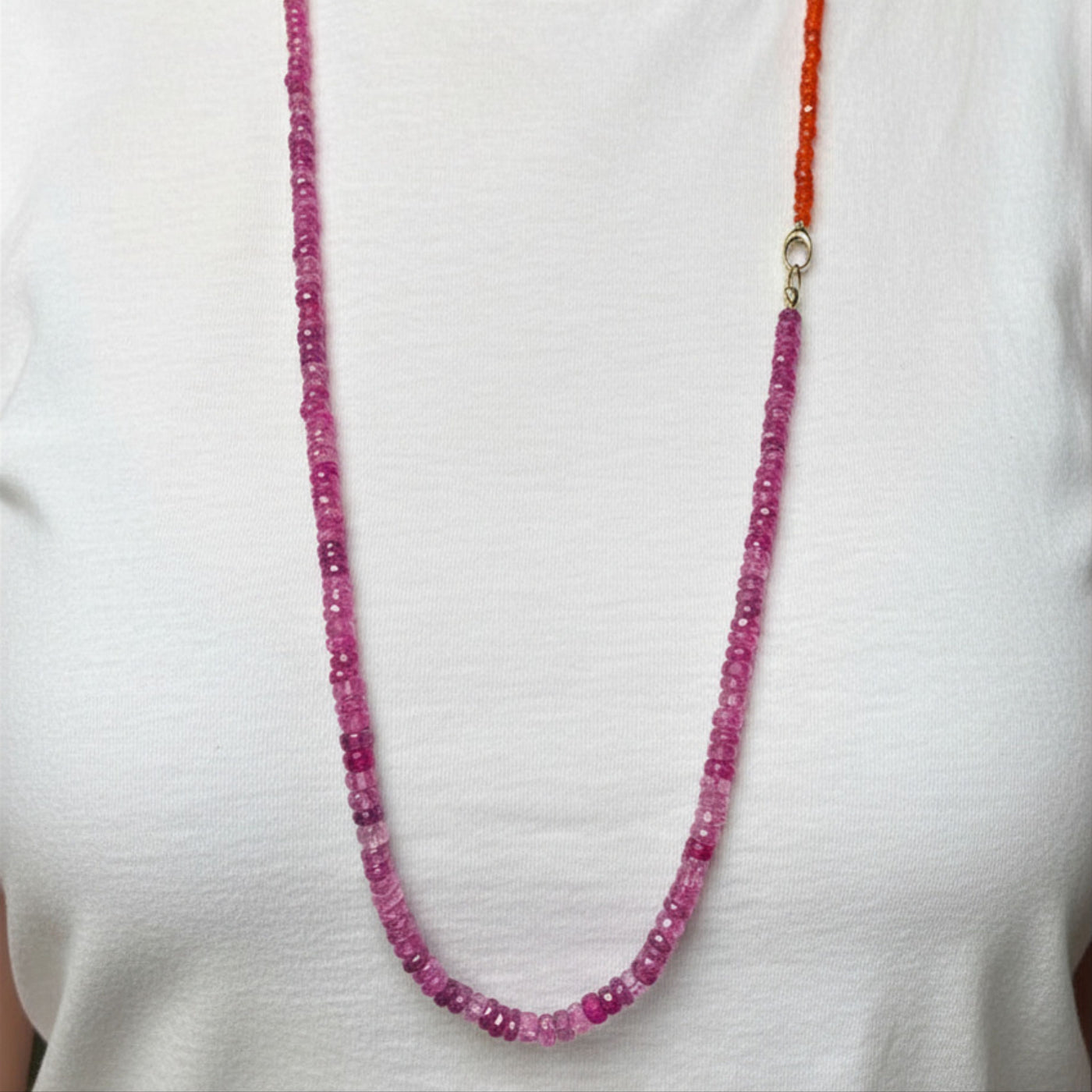 A woman in a plain white t-shirt shows how you can use your AMANDA PEARL evolution clasp to attach a fuchsia sapphire necklace to a carnelian faceted necklace to make one long, two tone necklace.