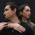 Two people standing close together with one person's hand on the other's shoulder, against a dark background. Both are wearing sterling silver rings, necklaces, and earrings.