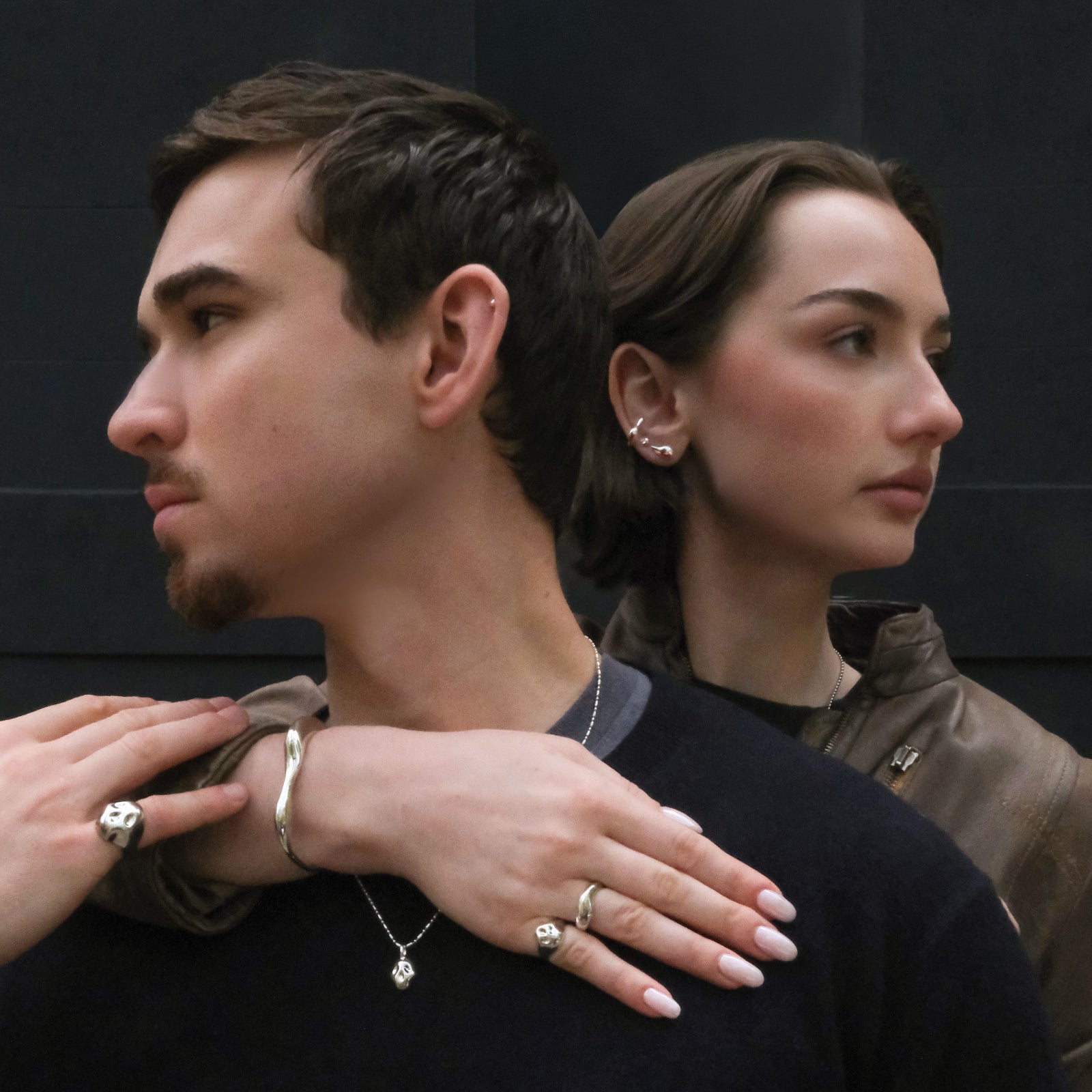 Two people standing close together with one person's hand on the other's shoulder, against a dark background. Both are wearing sterling silver rings, necklaces, and earrings.
