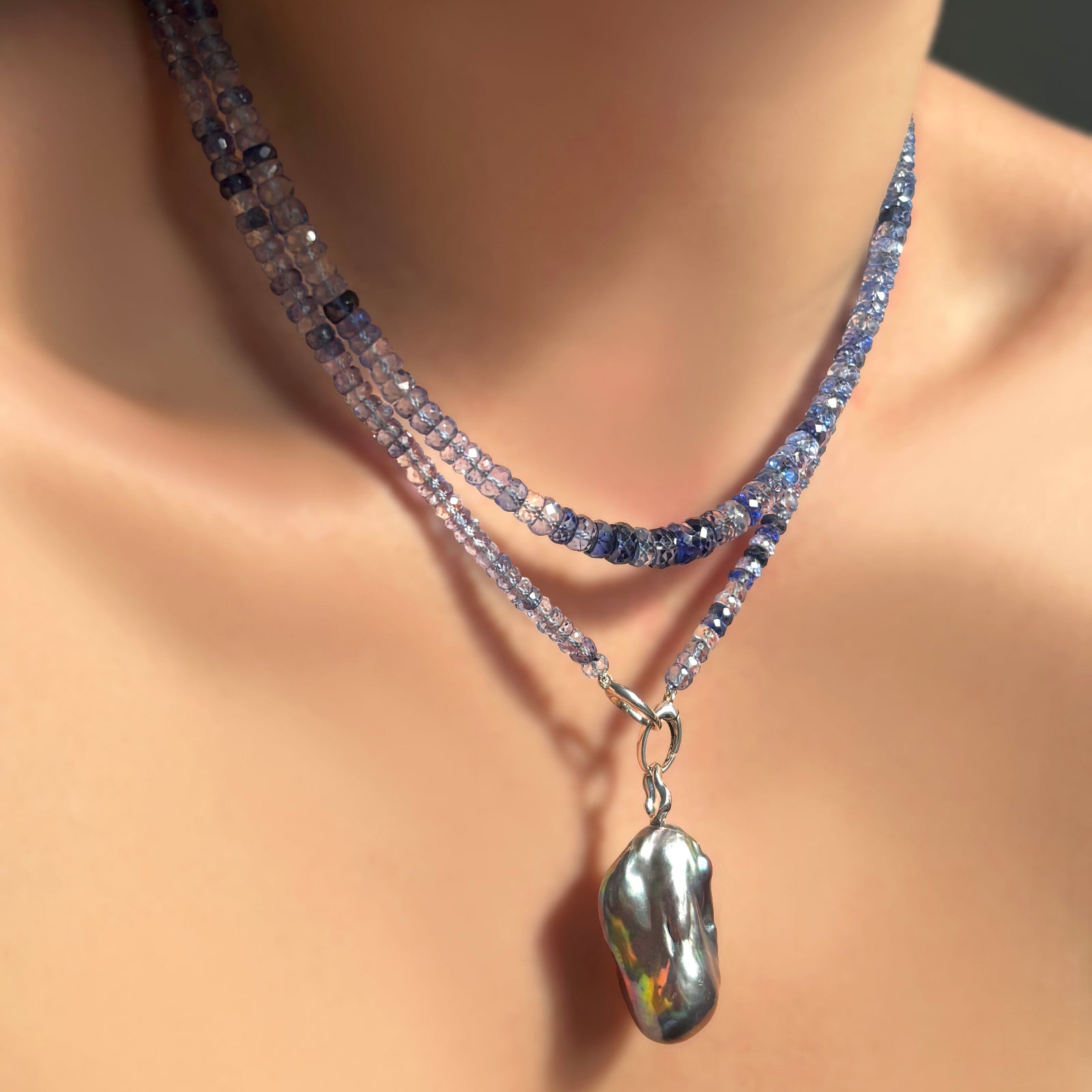 denim blue faceted sapphire necklaces with white gold linking ovals clasp dropping a silvery lunar baroque pearl charm on ripple bale on a model's neck.