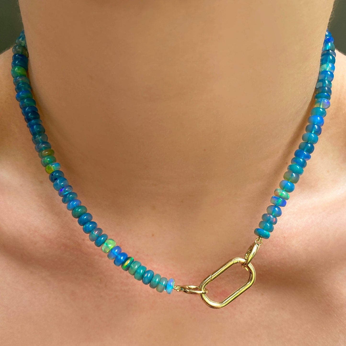 Shimmering beaded necklace made of smooth opal rondels in shades of blue, teal, and green on a slim gold oval clasp. Styled on a neck layered with plain oval charm lock.