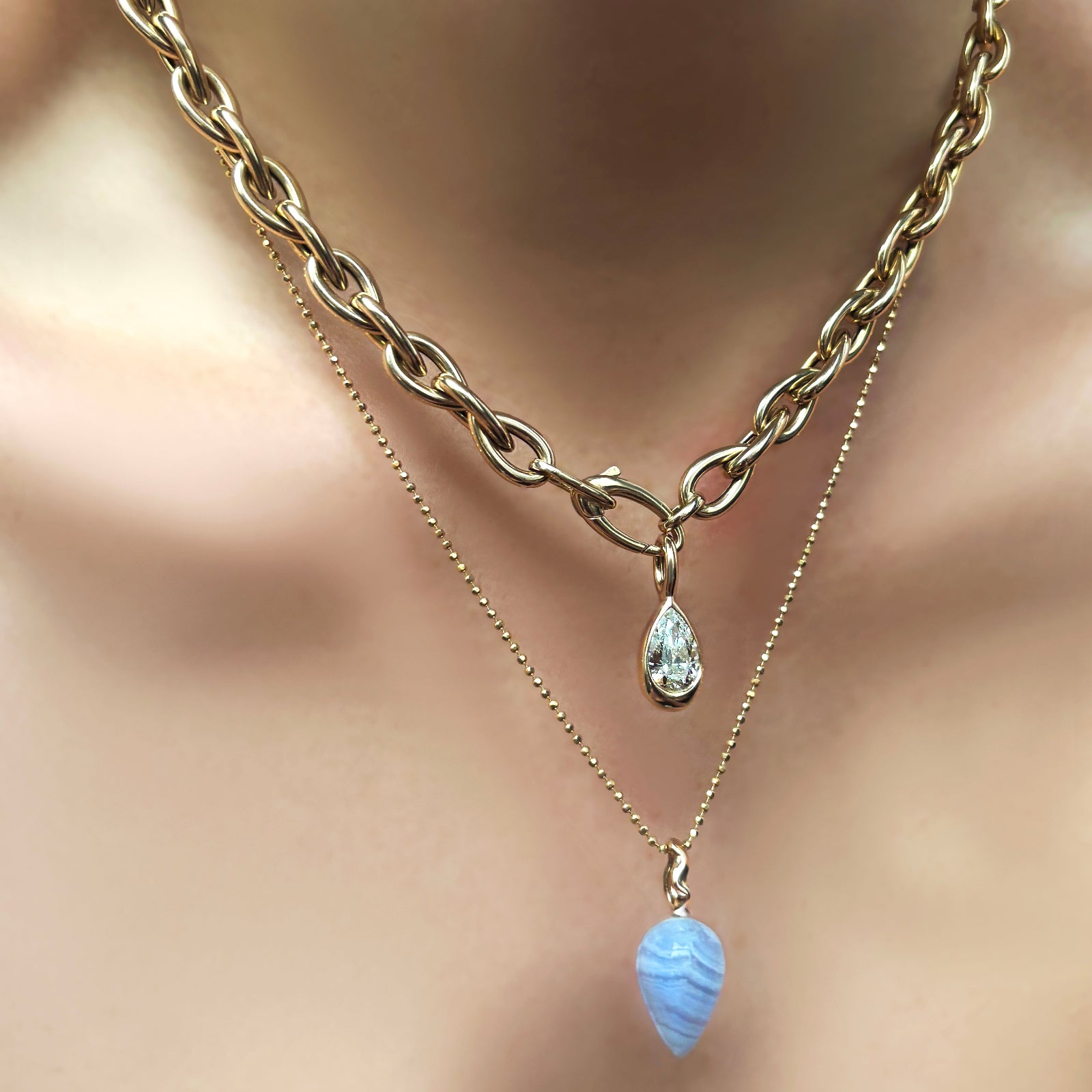 A model wears a drop link chain in 14k yellow gold from which hangs a diamond pear charm with a yellow gold bezel. A diamond cut bead chain hangs around the models neck as well and off of it hangs a blue lace agate gemstone drop charm. 