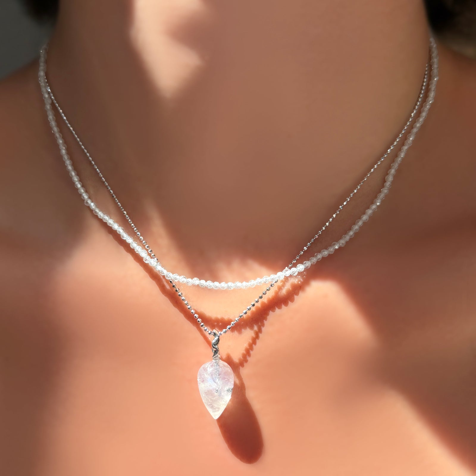 An AMANDA PEARL White Zircon slim gemstone necklace layered over a white gold diamond cut bead chain with rainbow moonstone drop charm on a model's neck.