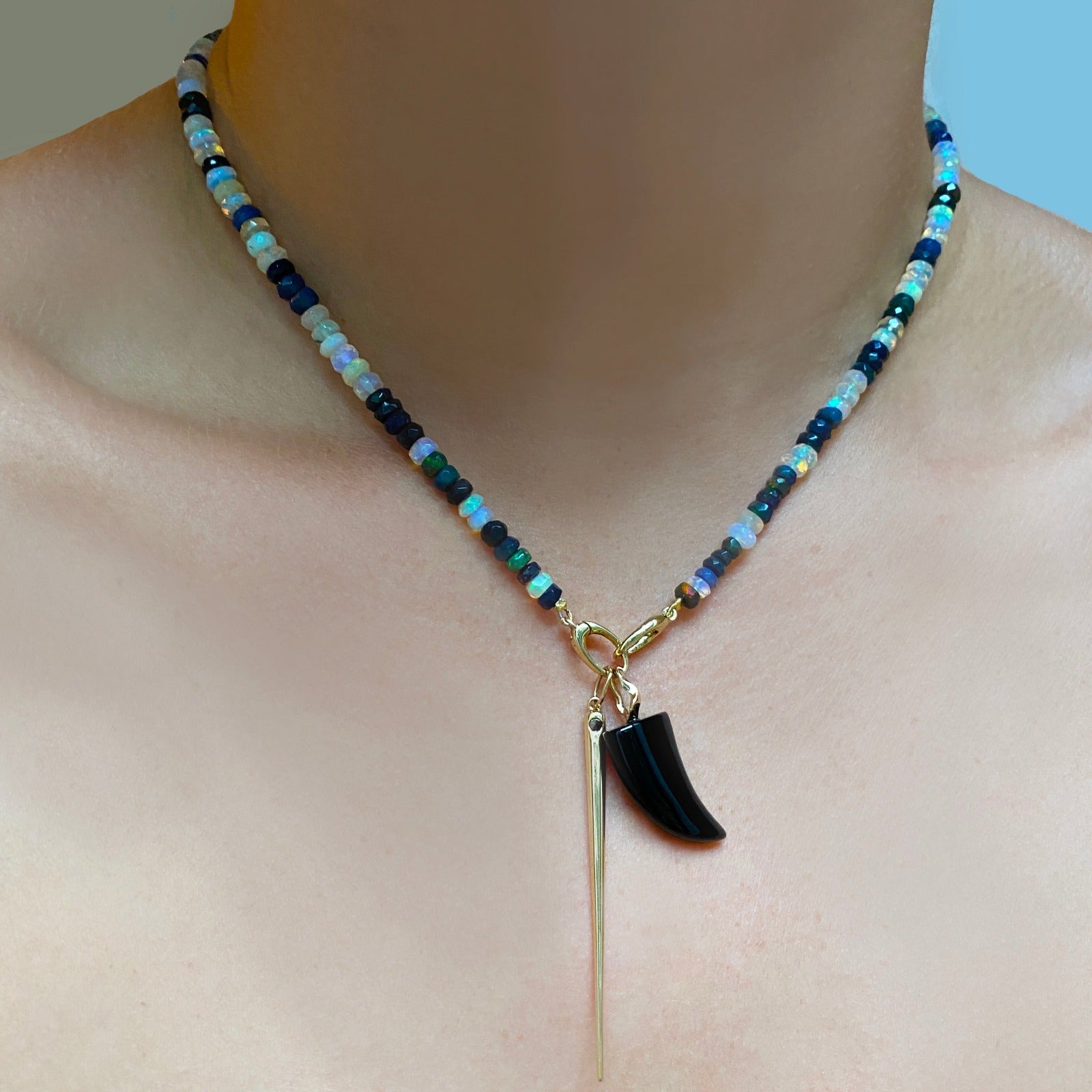 Shimmering beaded necklace made of faceted opals in shades of fiery pastels, grey, teal, and black on a gold linking ovals clasp. Styled on a neck layered with a horn and quill spike charm. 