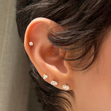 Close-up of an ear wearing AMANDA PEARL Elements diamond stud earrings with round, trillion, with a neutral background.val, and pear-shaped diamonds against a neutral background.