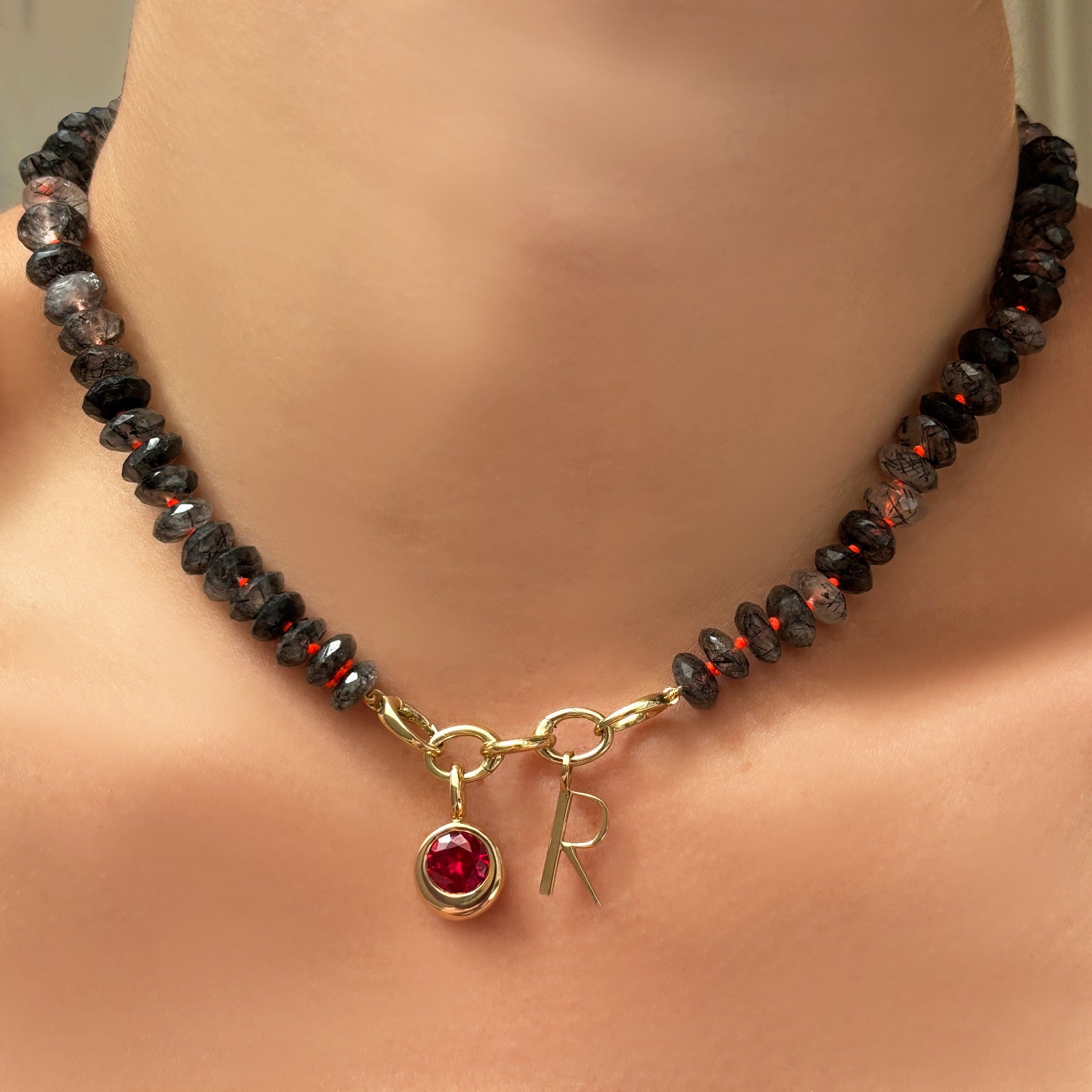 faceted rutilated quartz collar with triple link charm lock ,round molten solitaire charm with ruby, and letter charm on a model's neck.