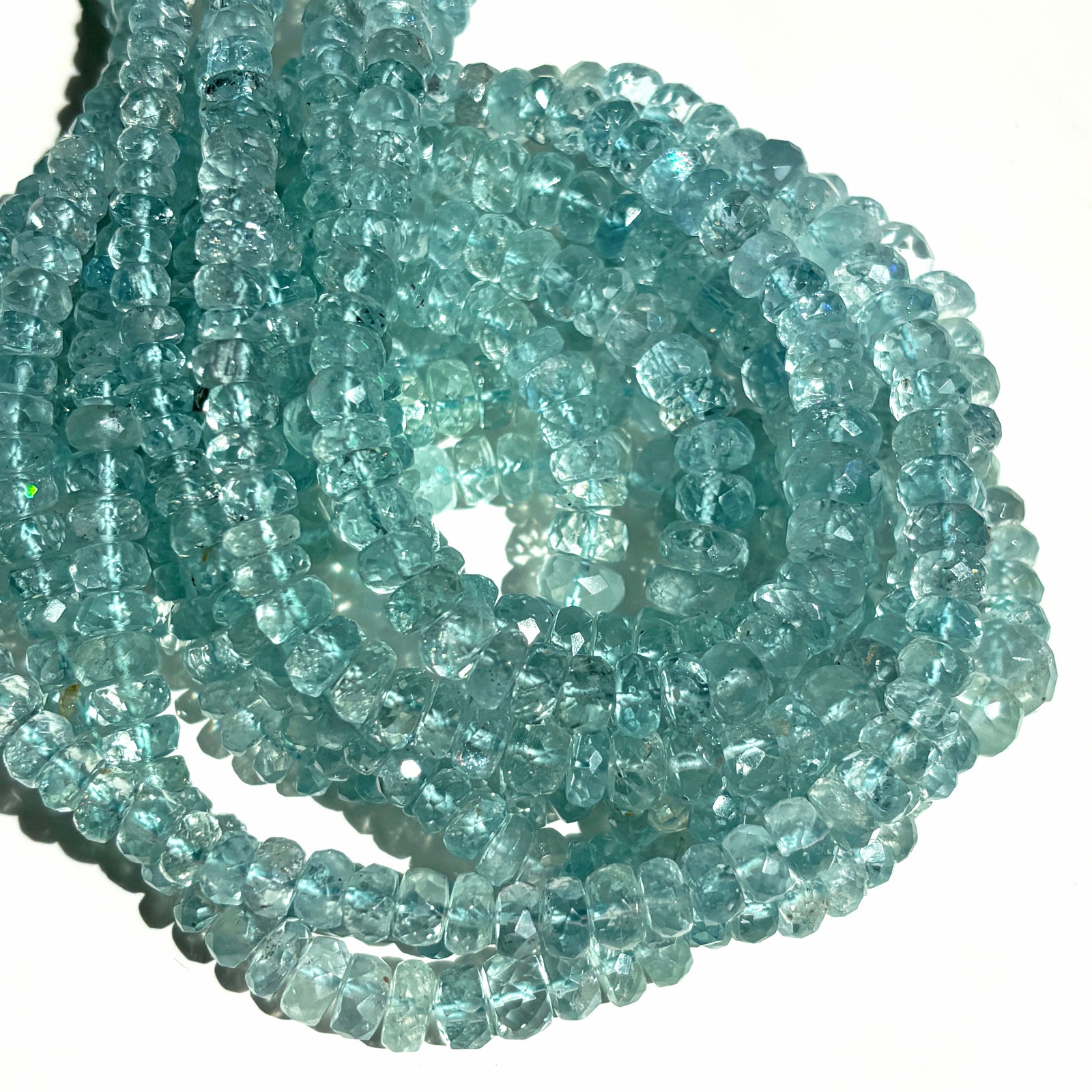 A pile of faceted aquamarine rondelle necklaces in sunlight on a white background.