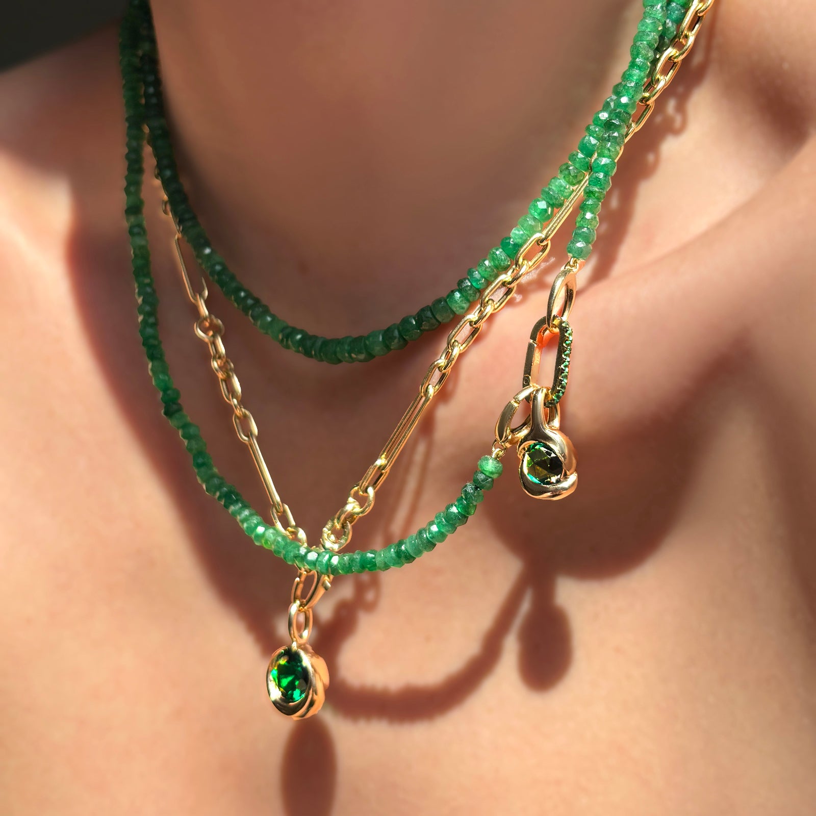 Faceted sapphire necklaces layered with irregular cable chan holding a molten solitaire charm with emerald, molten knot charm with rose cut emerald, and small emerald charm lock on a model's neck.
