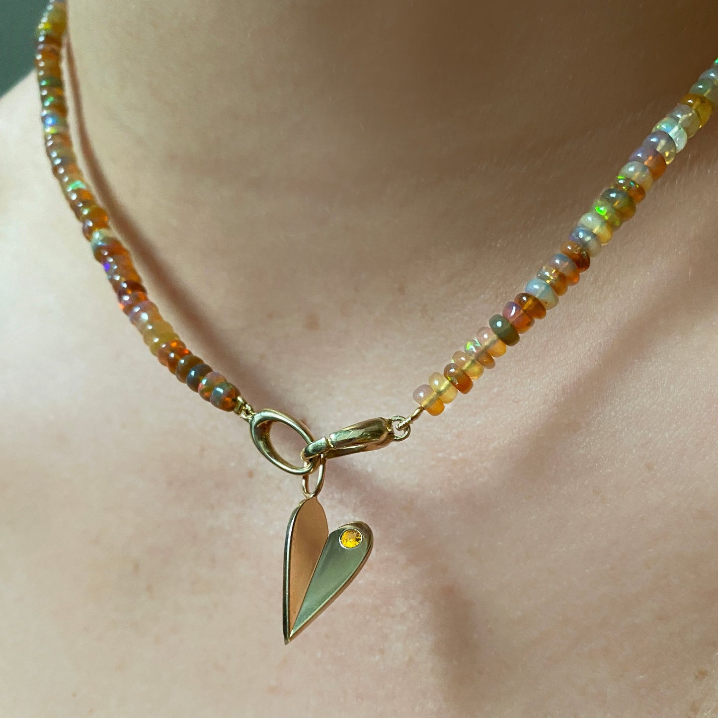 Shimmering beaded necklace made of smooth opal rondels in shades of light yellow, light orange, white, and brown on a slim gold lobster clasp. Styled on a neck layered with a folded heart charm.
