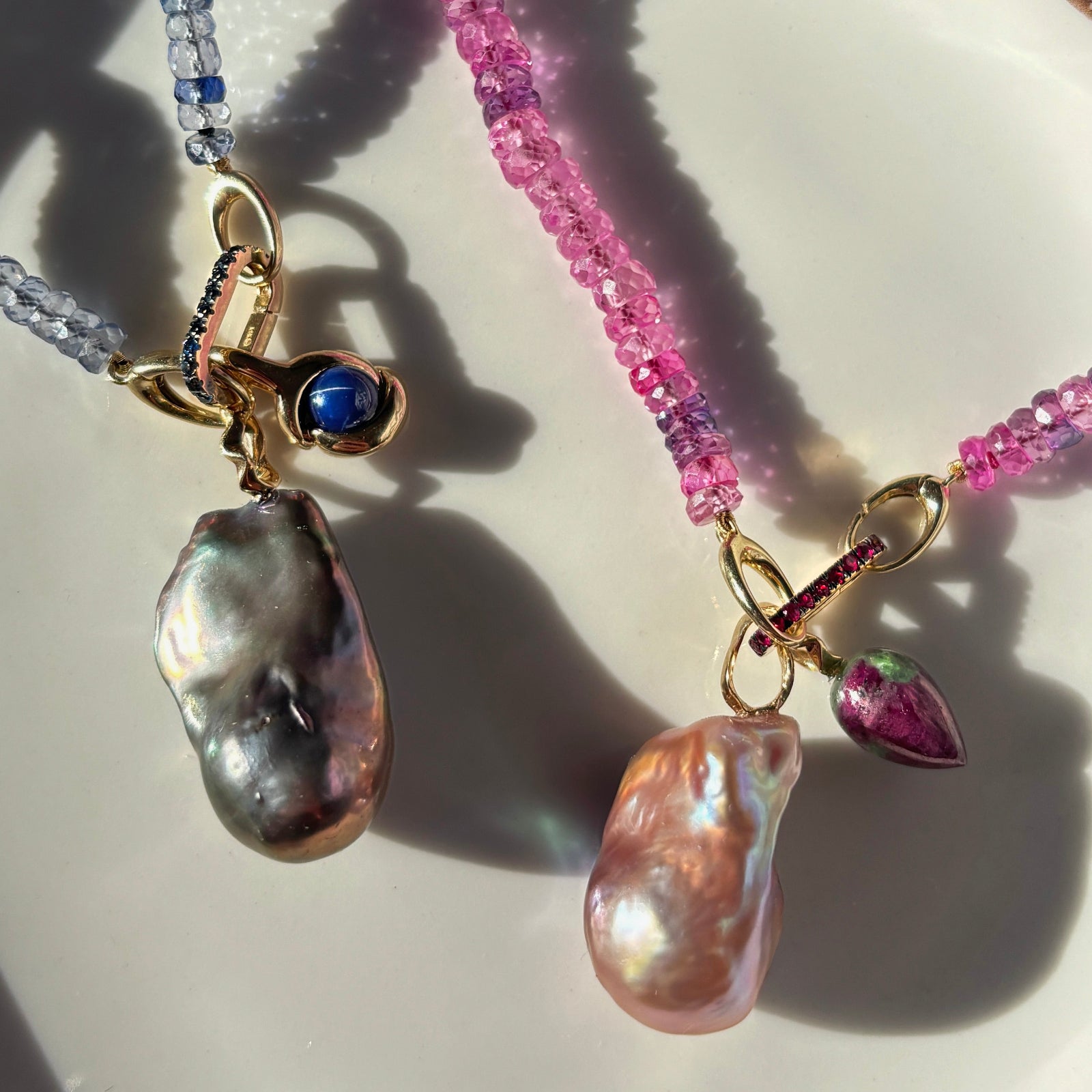 Denim blue faceted sapphire necklace with sapphire charm lock and star sapphire molten knot charm next to a fuchsia faceted sapphire necklace with ruby charm lock, blush baroque pearl charm and ruby zoisite drop charm. 