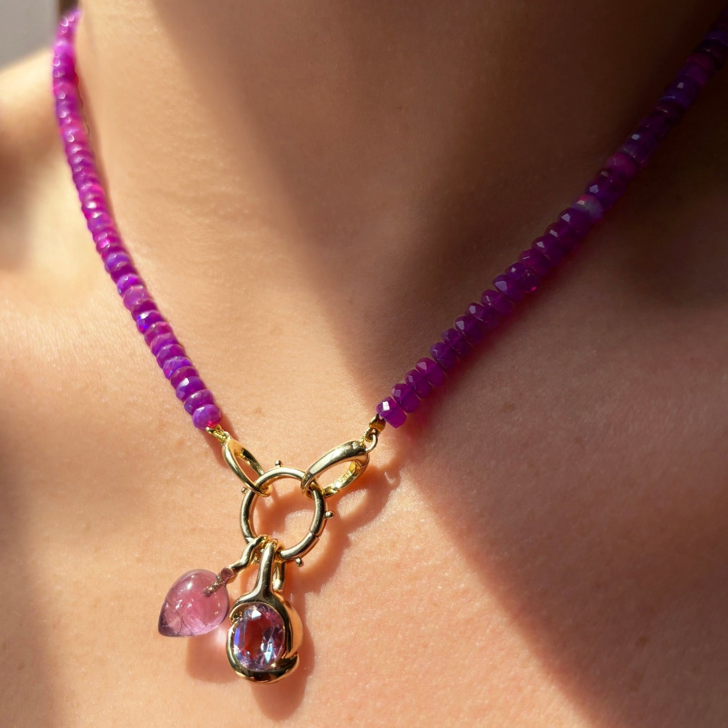 Shimmering beaded necklace made of faceted opals in shades of fuchsia pink on a gold linking ovals clasp. Styled on a neck layered with the beaded round charm lock, amethysts molten knot charm and acorn charm. 