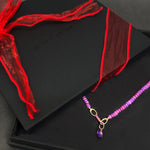 Black box with a red ribbon and a purple beaded necklace on a dark surface.