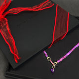 Black box with a red ribbon and a purple beaded necklace on a dark surface.
