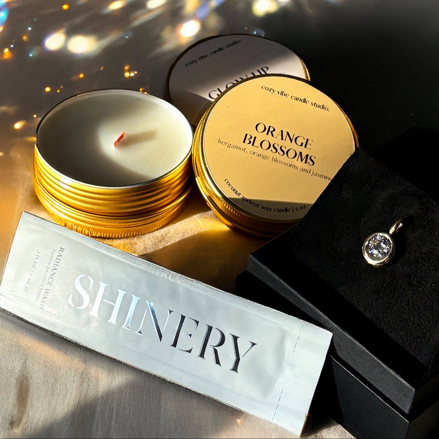 Beautifully packaged candle, shinery jewelry wash sample, next to gold and diamond charm with sparkle on the wall behind.
