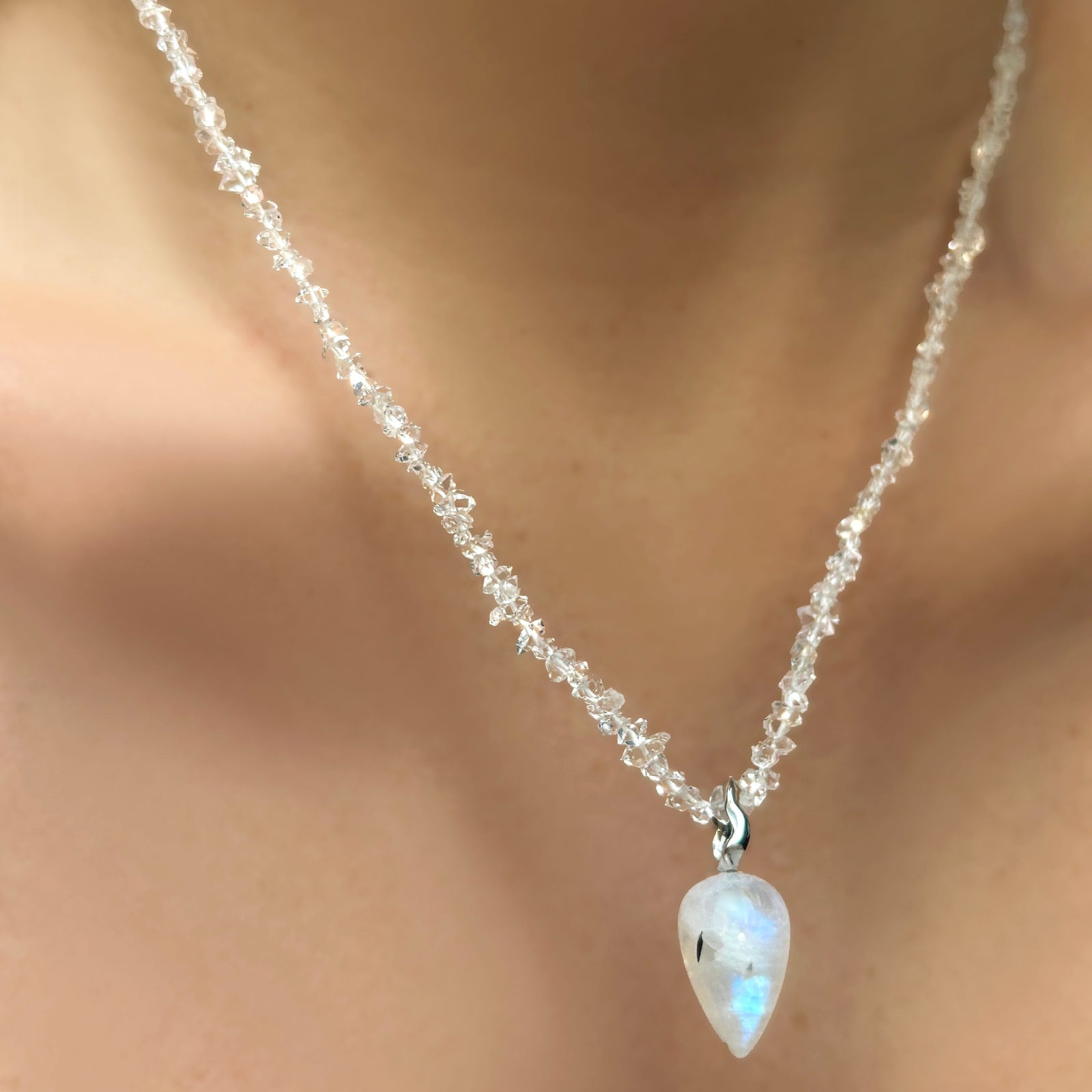A herkimer necklace hangs on a model's neck. From it is strung a moonstone drop charm with a 14K recycled white gold moonstone drop charm.