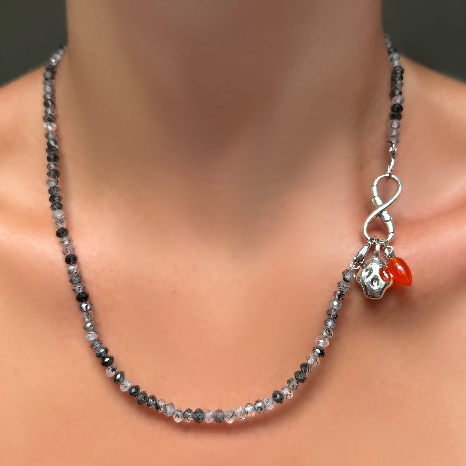 Faceted Rutilated Quartz Necklace with recycled sterling silver molten infinity charm lock holing a molten skull charm and carnelian drop charm with silver ripple bale on a model's neck.