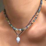 A model wears, a lace agate drop charm with a recycled 14K white gold ripple bale. It hangs from an iolite slim gemstone necklace, and is coupled with a galaxy faceted opal necklace. 