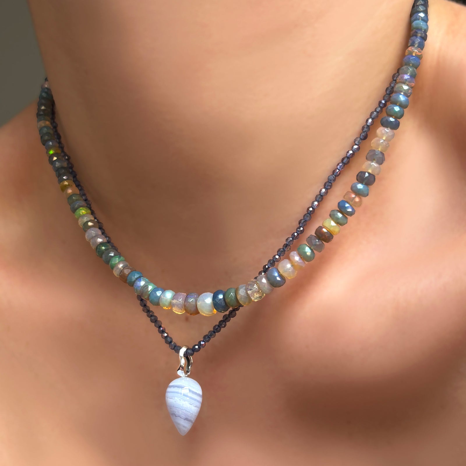 A model wears, a lace agate drop charm with a recycled 14K white gold ripple bale. It hangs from an iolite slim gemstone necklace, and is coupled with a galaxy faceted opal necklace. 