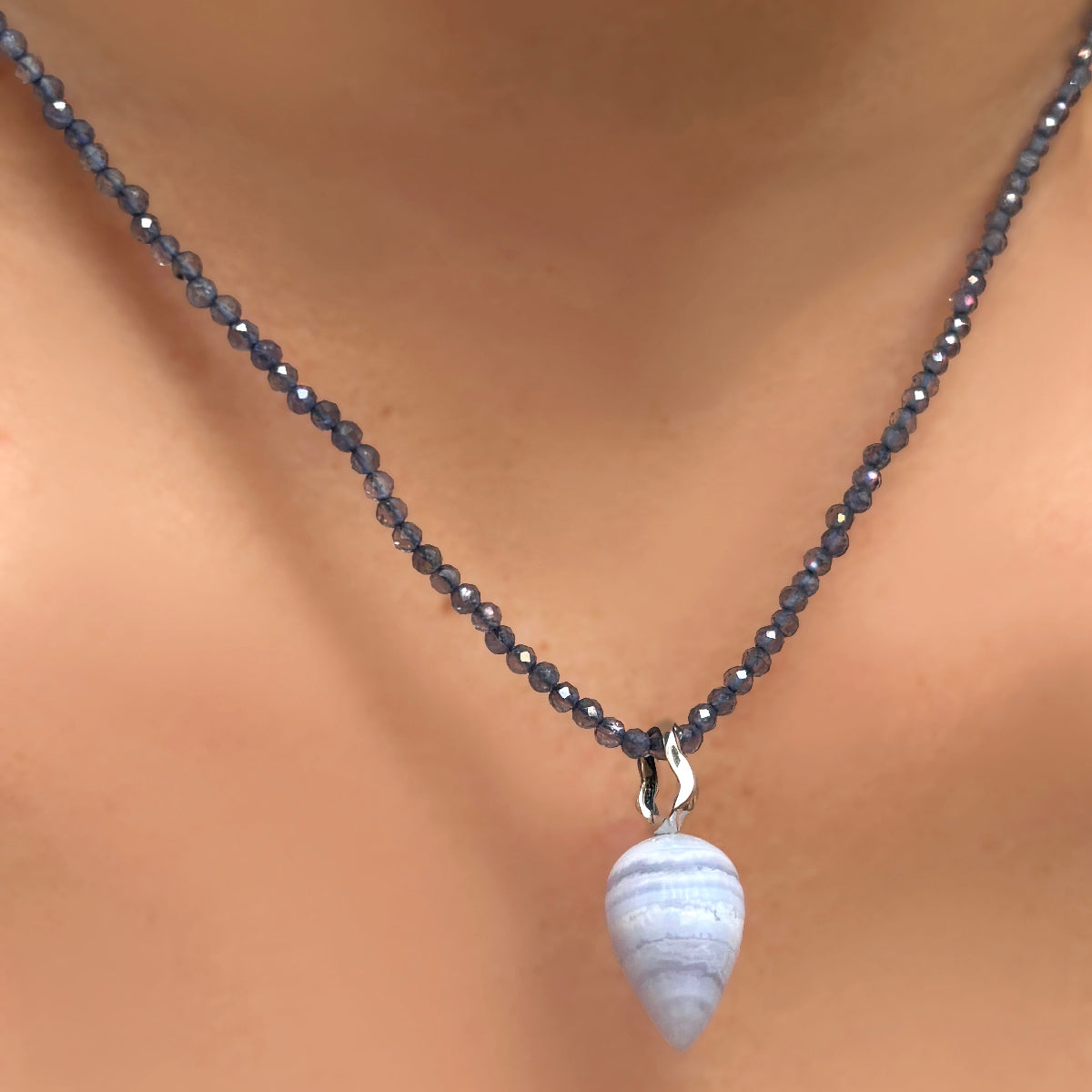 A close-up of lace agate charm drop with a recycled white gold 14K bale hangs on a deep grey purple iolite necklace with faceted sparkling beads.