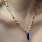 A model wears an irregular paperclip chain necklace in 14K yellow gold, from which hangs a long lapis drop charm with a 14K recycled yellow gold ripple bale. 