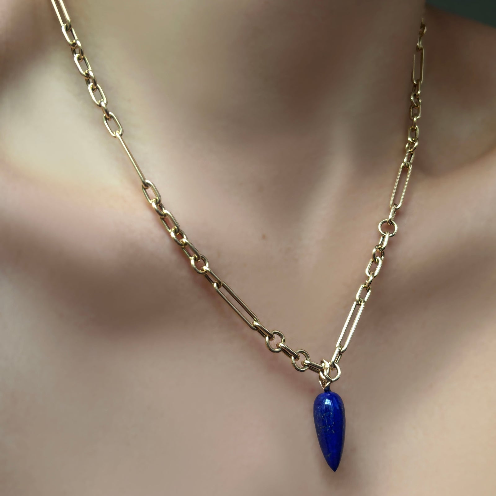 A model wears an irregular paperclip chain necklace in 14K yellow gold, from which hangs a long lapis drop charm with a 14K recycled yellow gold ripple bale. 