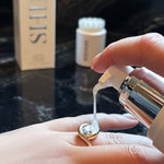 Hand applying a dollop of Shinery jewelry wash to a gold and diamond AMANDA PEARL ring with a blurred background