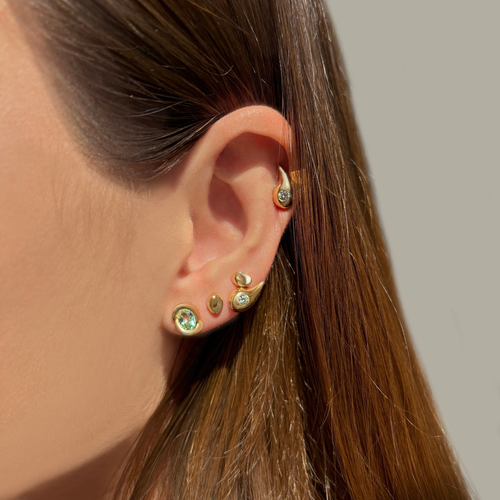 A Person wearing various shaped earrings. Including a gemstone stud, yellow gold studs and teardrop-shaped studs with diamond accent