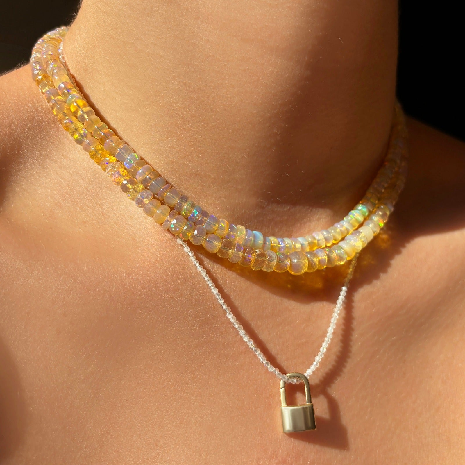 Shimmering beaded necklace made of faceted opals in shades of clear and light yellow on a gold linking ovals clasp. Styled on a neck layered with a padlock charm.