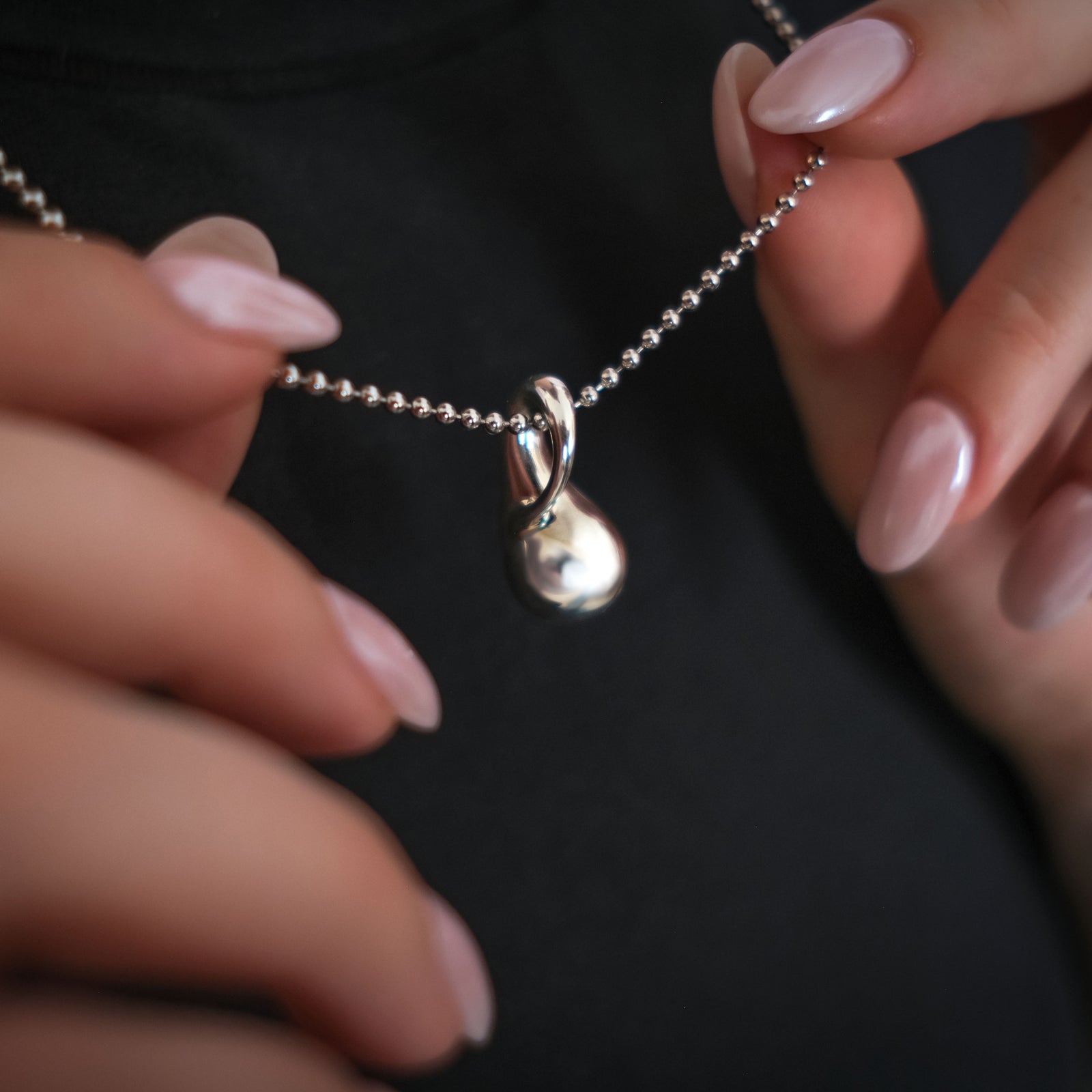 AMANDA PEARL Sterling silver bead chain necklace with a molten drop charm held by hands against a dark background
