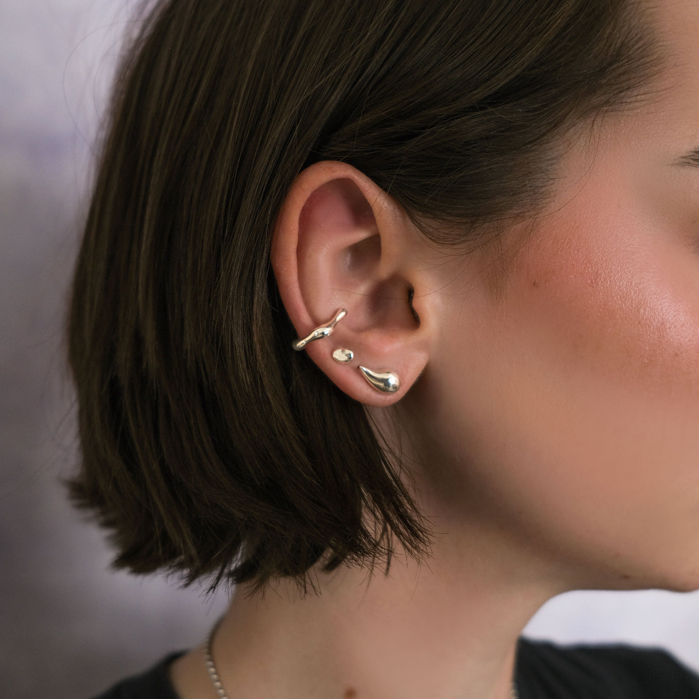A woman with a dark bob in a black t shirt wears her AMANDA PEARL molten earring, small molten stud, and a molten ear cuff. 