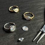 Three molten solitaire rings lay with a pair of pliers and two loose diamonds. The round ring is white gold, and the marquise and pear are yellow gold. They lay on a grey wood grain background.