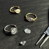 Three molten solitaire rings lay with a pair of pliers and two loose diamonds. The round ring is white gold, and the marquise and pear are yellow gold. They lay on a grey wood grain background.