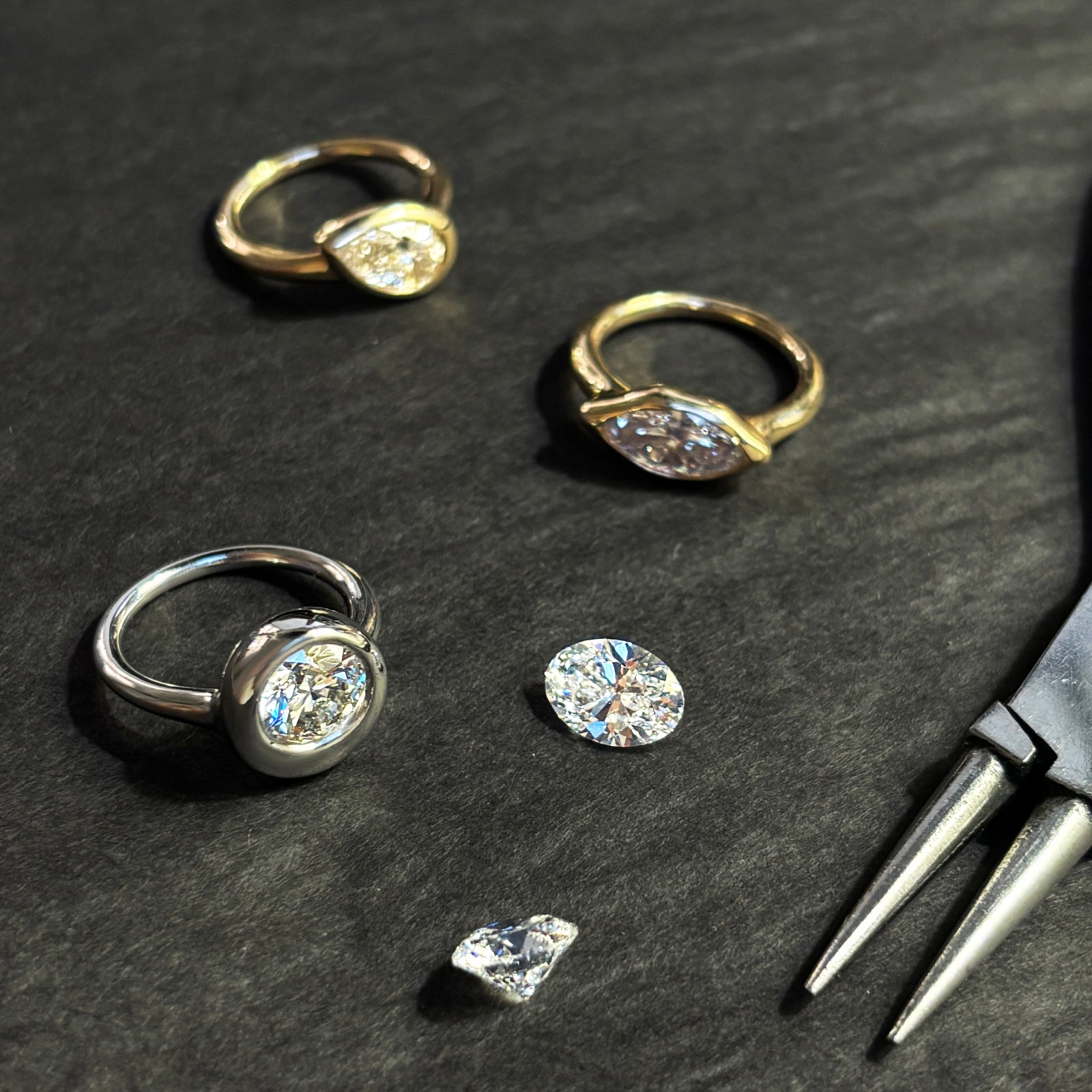 Three molten solitaire rings lay with a pair of pliers and two loose diamonds. The round ring is white gold, and the marquise and pear are yellow gold. They lay on a grey wood grain background.