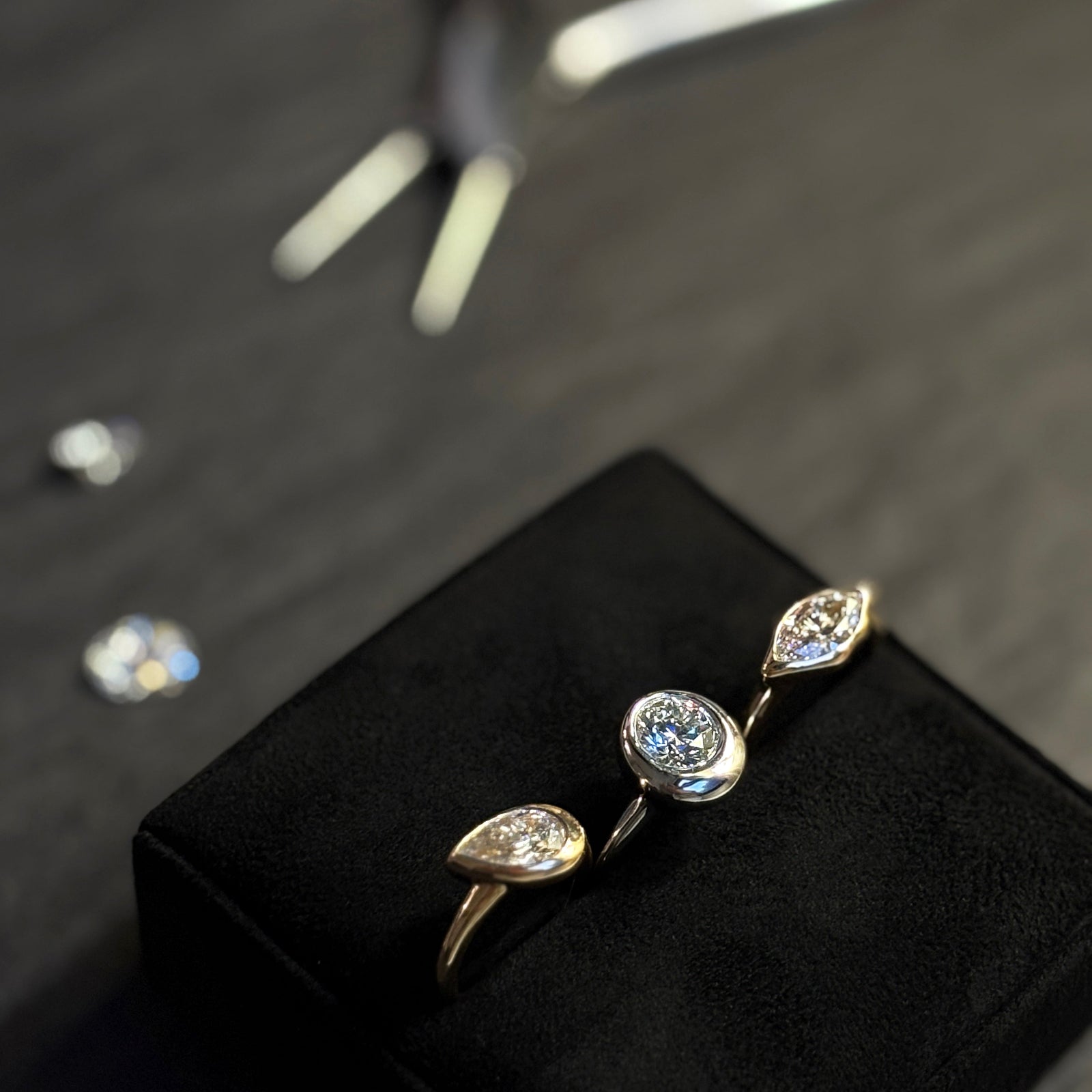 Three molten solitaire rings rest on a jewelry pillow. The round and marquise rings are white gold, and the pear is yellow gold. In the Background rests a pair of jewelry pliers and some loose diamonds. The rings feature 14K recycled gold and ethically sourced lab grown diamonds.
