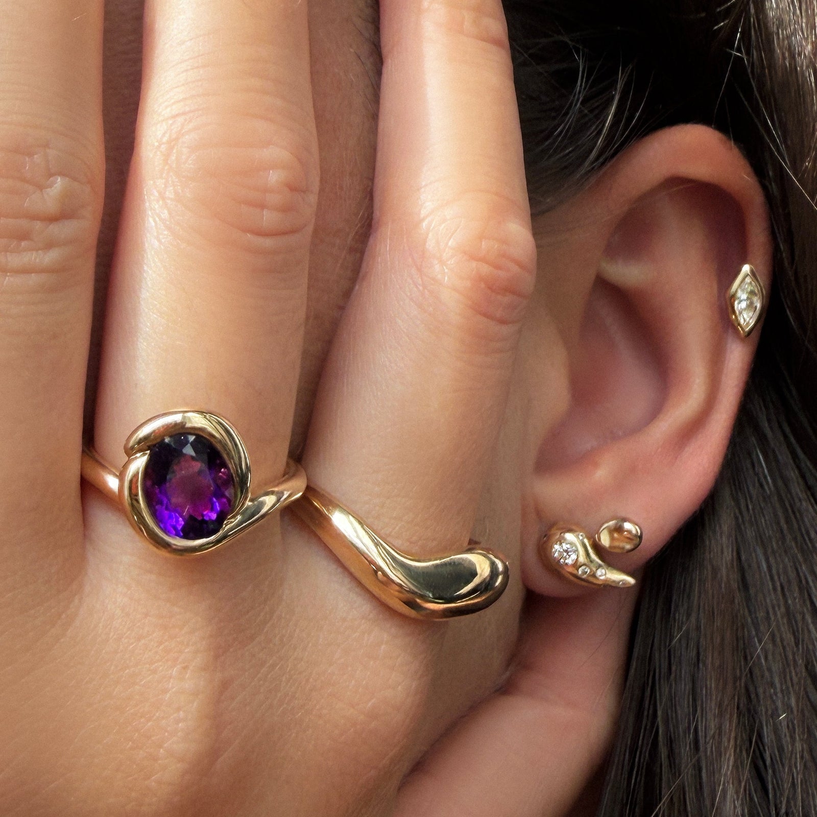 AMANDA PEARL recycled 14k gold knot ring with deep purple amethyst worn on a model's hand with a gold molten ring, next to an ear with the molten studs with diamonds, small molten stud, and molten marquise solitaire stud.