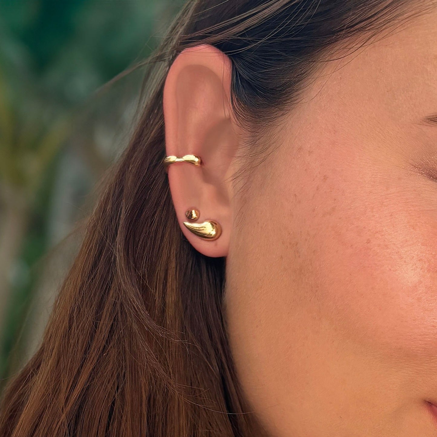 Small liquid round yellow gold stud earring on ear, styled with multiple gold earrings