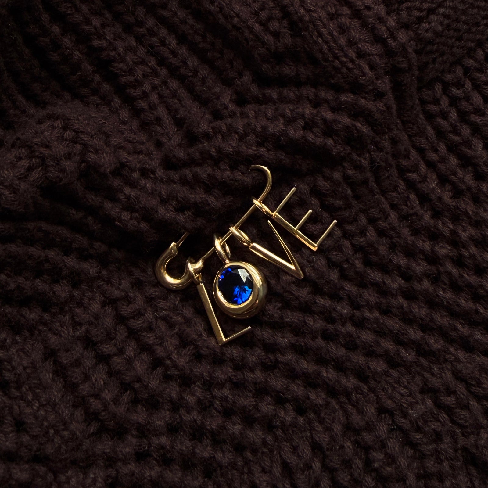 A recycled 14k gold molten safety pin iin a dark chunky knit sweater, dropping letter charms and a round molten sapphire solitaire charm spelling out LOVE.