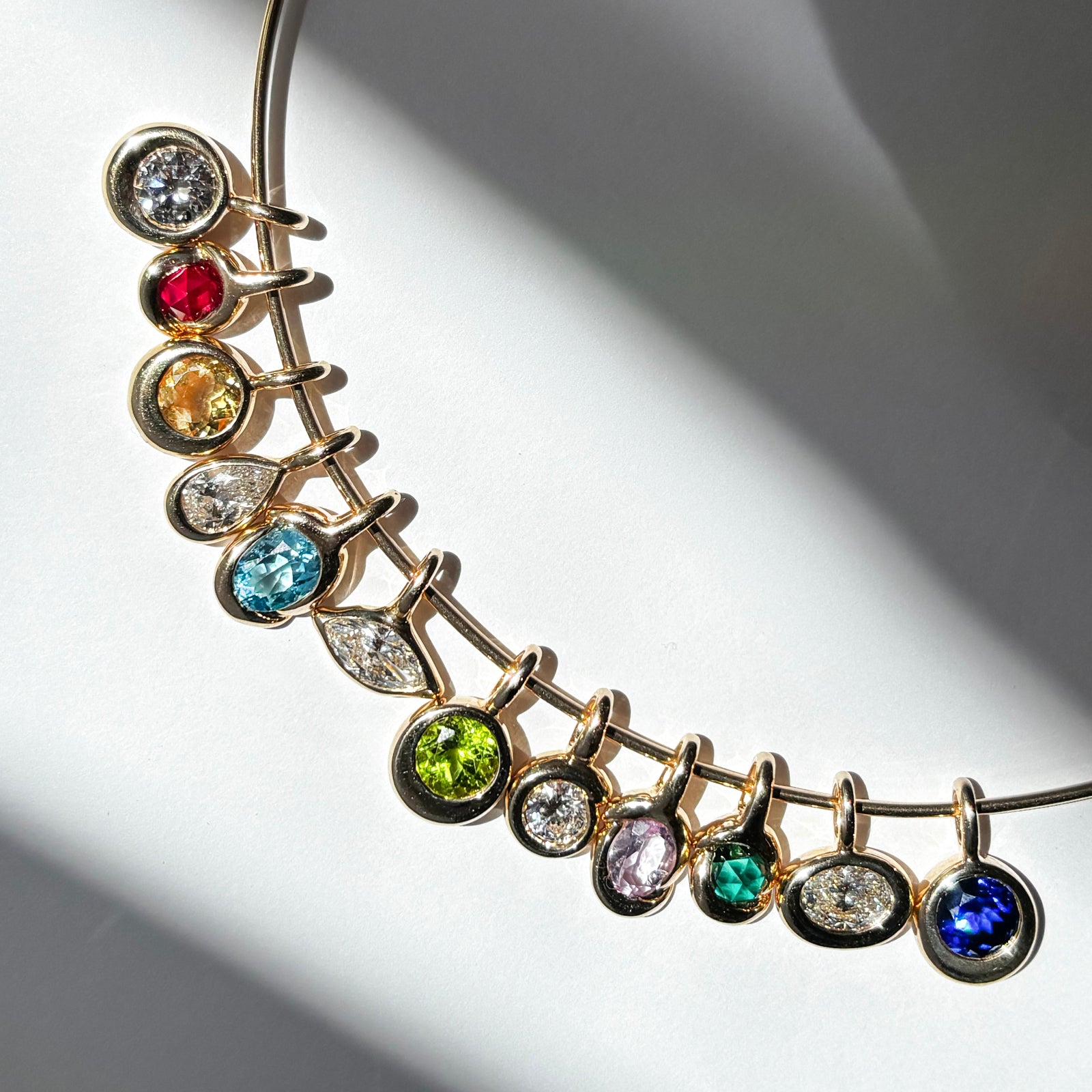 AMANDA PEARL Wire choker with an assortment of molten solitaire charms with diamonds and colored gemstones on a white background framed by shadows.